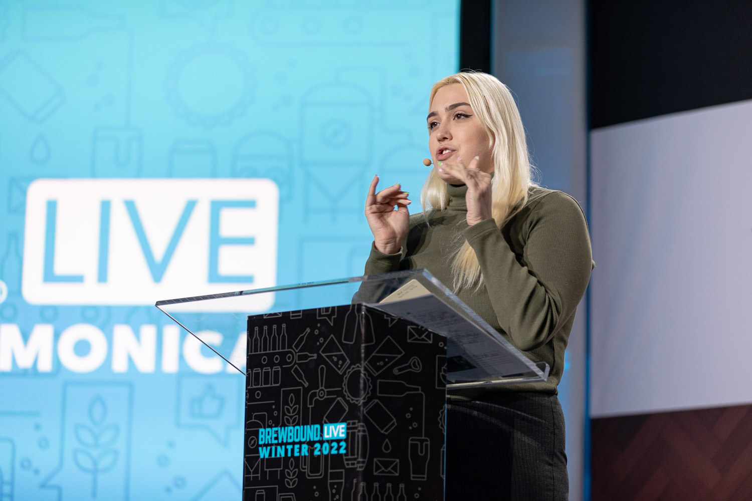 A woman with long blonde hair gestures while speaking at a clear podium on a blue stage with a Brewbound Live Winter 2022 sign.