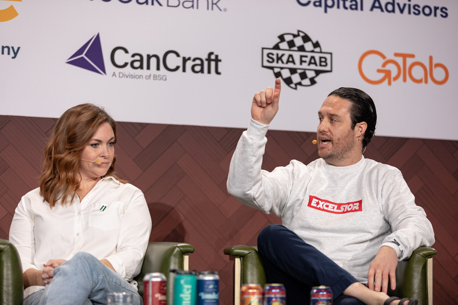 A man in a white Excelsior sweatshirt points upward while a woman beside him looks on, with cans on the table in front of them.