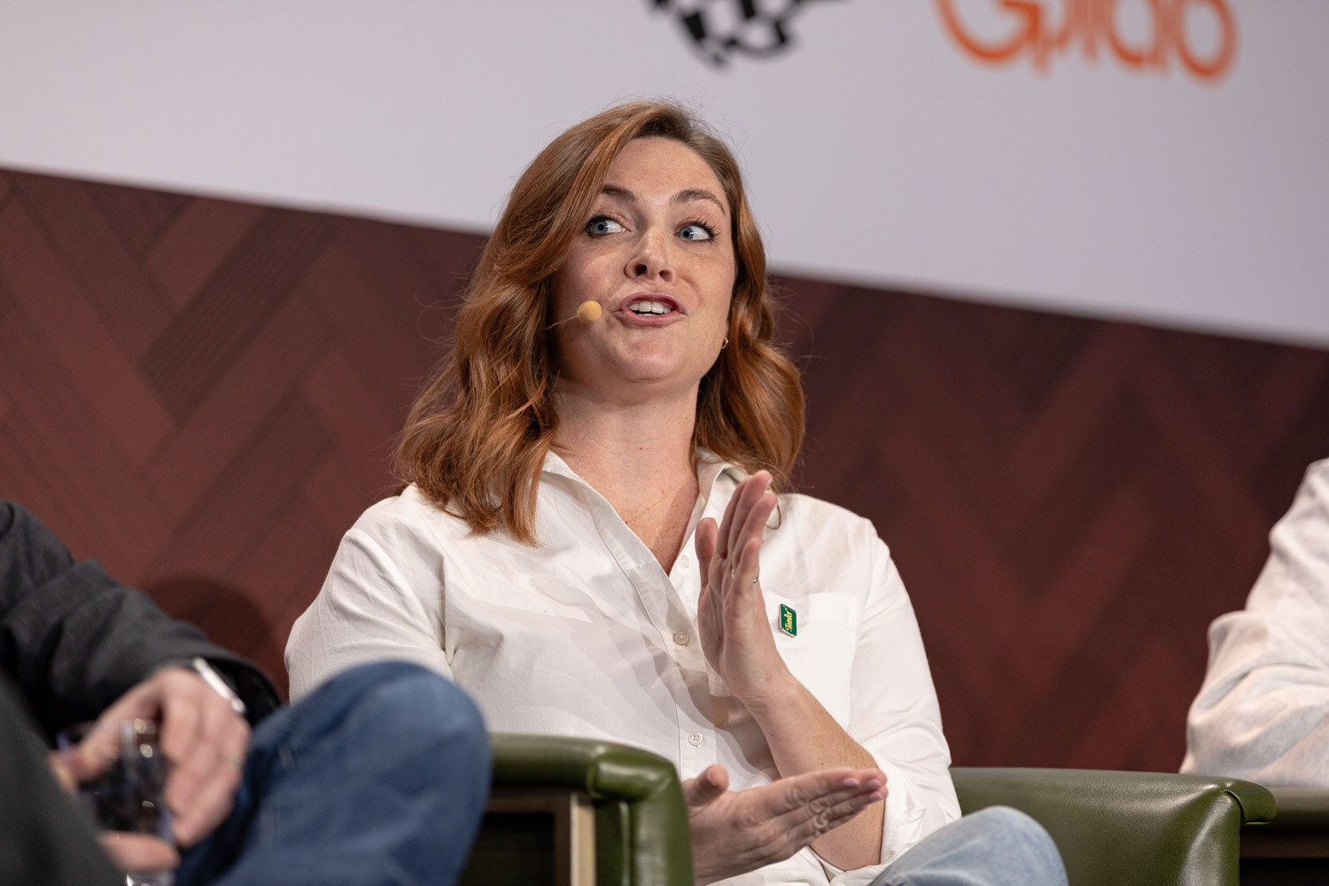 A woman with auburn hair in a white button-up shirt speaks and gestures with open hands on stage.
