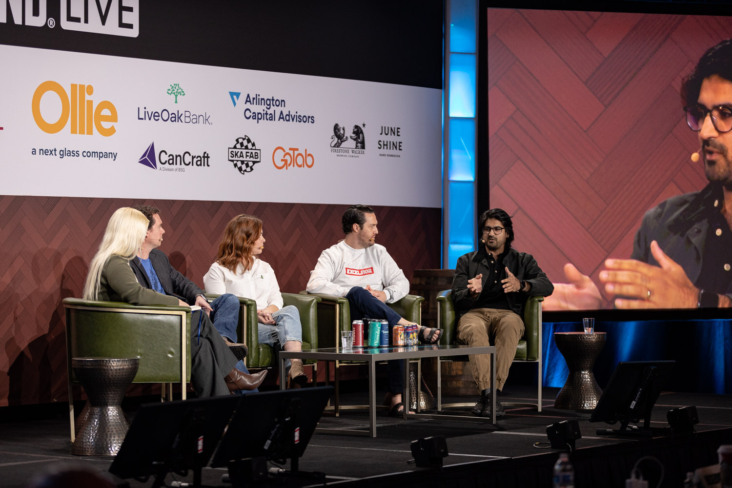 Wide view of five people seated on a stage in green armchairs with cans on a table, under a Brewbound Live banner with sponsor logos including Ollie, Live Oak Bank and CanCraft.