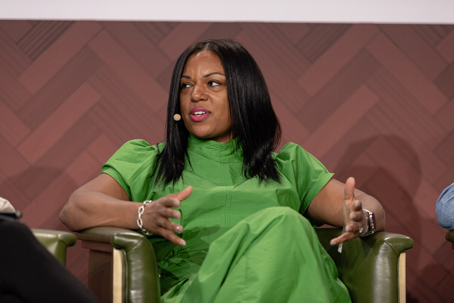 A woman with long dark hair in a bright green dress gestures with one hand while seated in a green armchair.