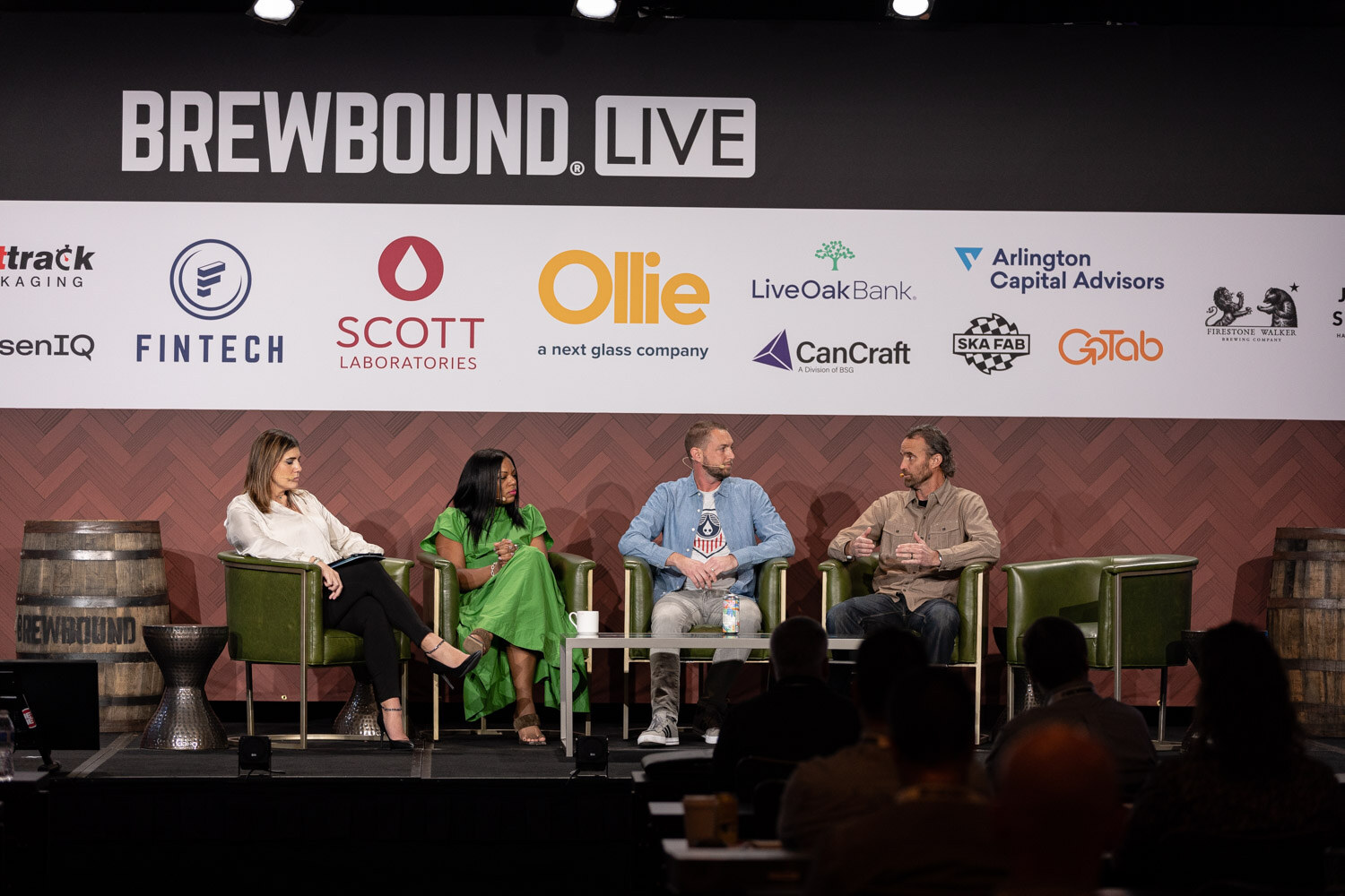 Wide view of four people in green armchairs on a stage with the Brewbound Live sponsor wall behind them and a barrel at left.