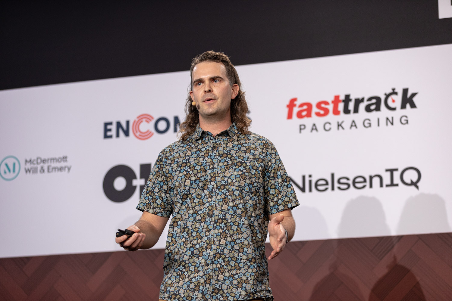 A man with shoulder-length hair in a patterned short-sleeve shirt holds a clicker in front of sponsor logos including Fasttrack Packaging and NielsenIQ.