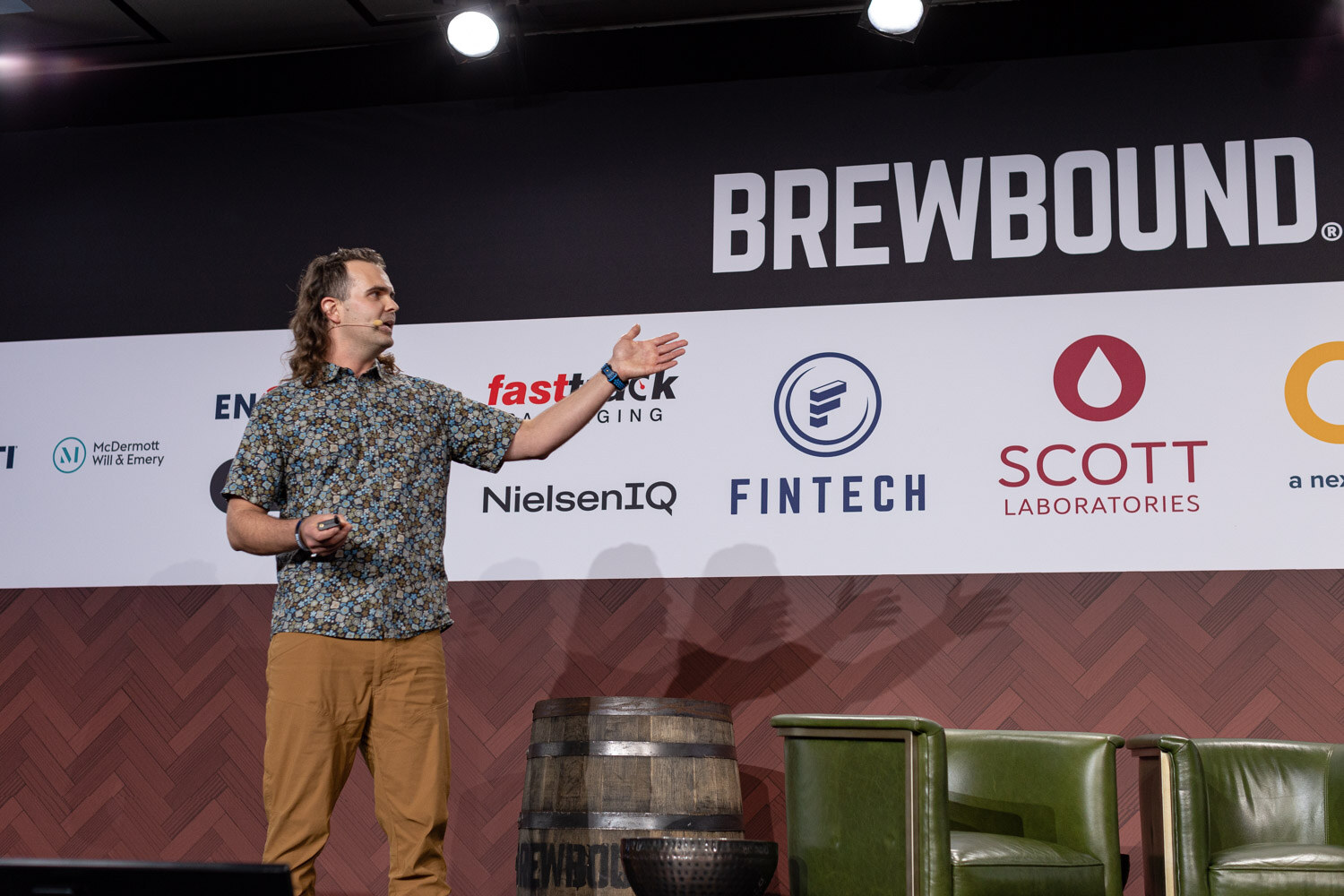A man in a patterned shirt stands on a stage with armchairs and a barrel behind him, one arm stretched out.