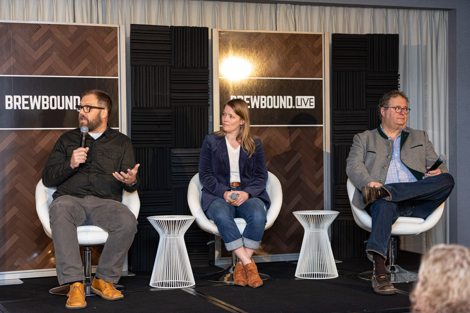 Wide view of three people seated in white chairs on a small stage in front of dark acoustic panels and Brewbound Live signs.