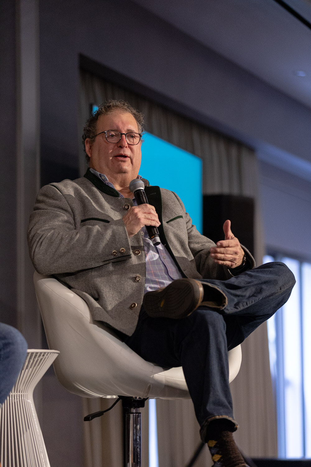 A man with glasses in a gray jacket and a blue patterned shirt holds a microphone while seated in a white chair on a small stage.