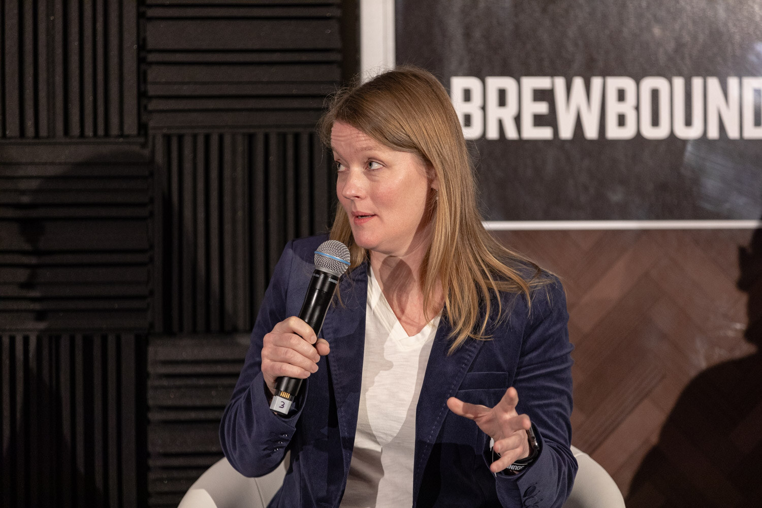 A woman with long auburn hair in a navy blazer holds a microphone and gestures in front of a Brewbound Live sign.
