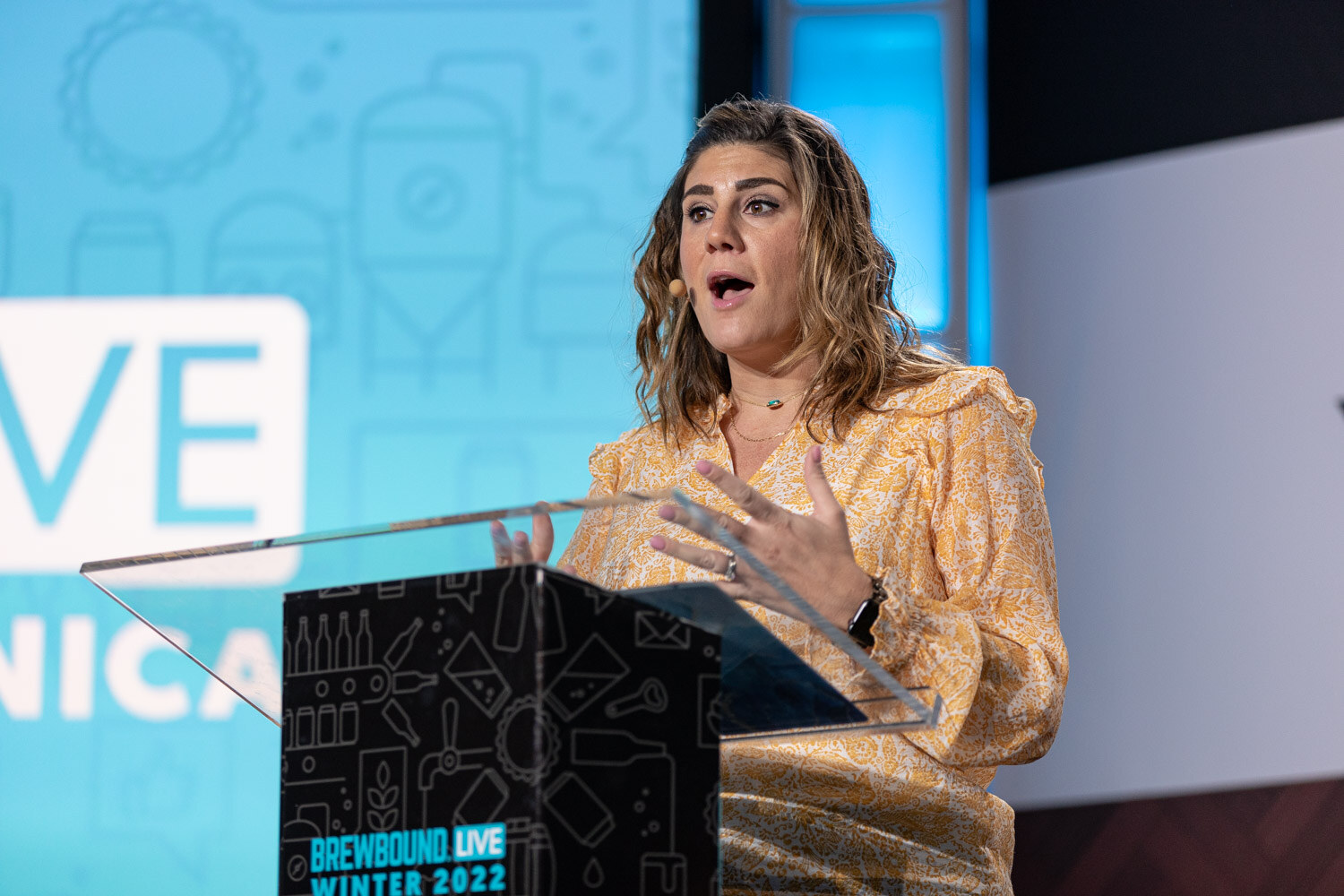 A woman with wavy brown hair in a yellow patterned blouse speaks at a clear podium in front of a blue Brewbound Live Santa Monica backdrop.
