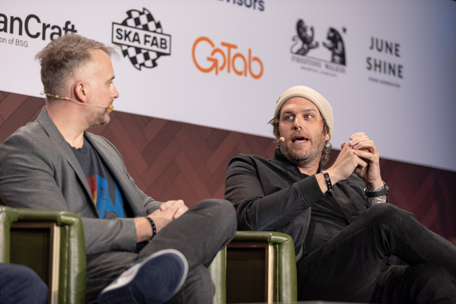 A man in a cream beanie and black jacket speaks with both hands raised while a man in a gray blazer listens.