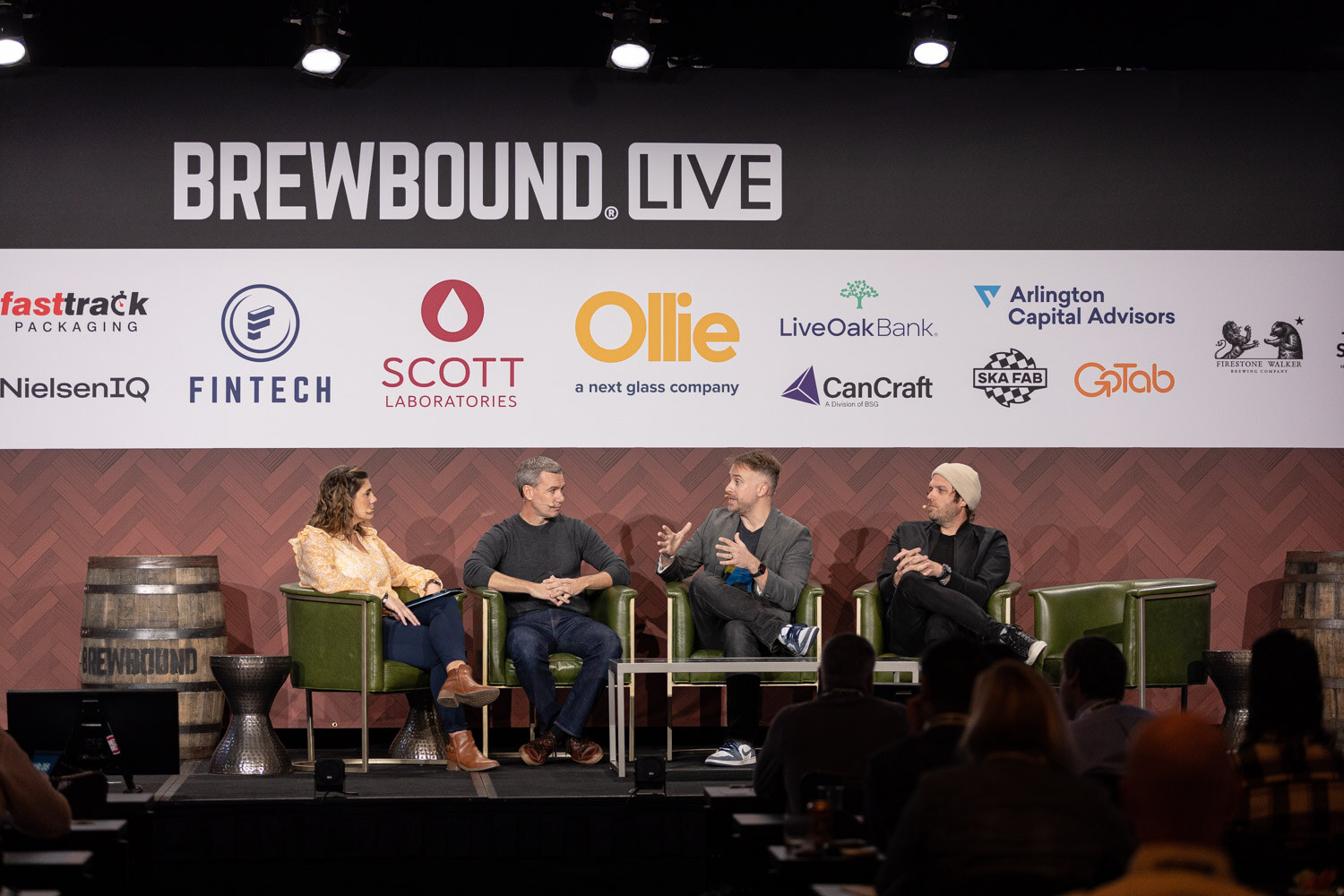 Wide view of four people seated in green armchairs on a stage under the Brewbound Live sponsor wall, with audience members in silhouette.