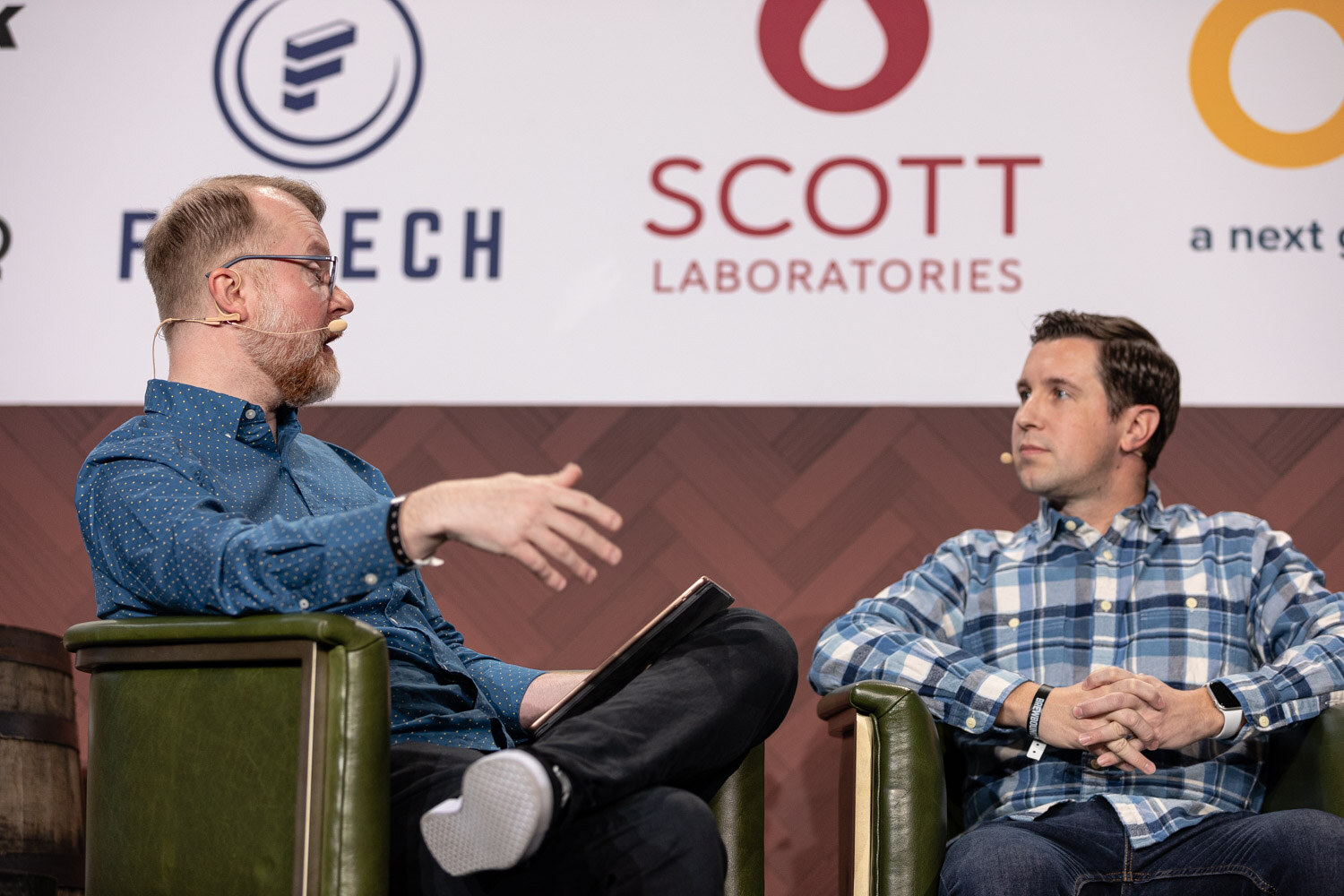A bearded man in a blue shirt holding a tablet gestures with one hand while a man in a plaid shirt sits beside him on stage.