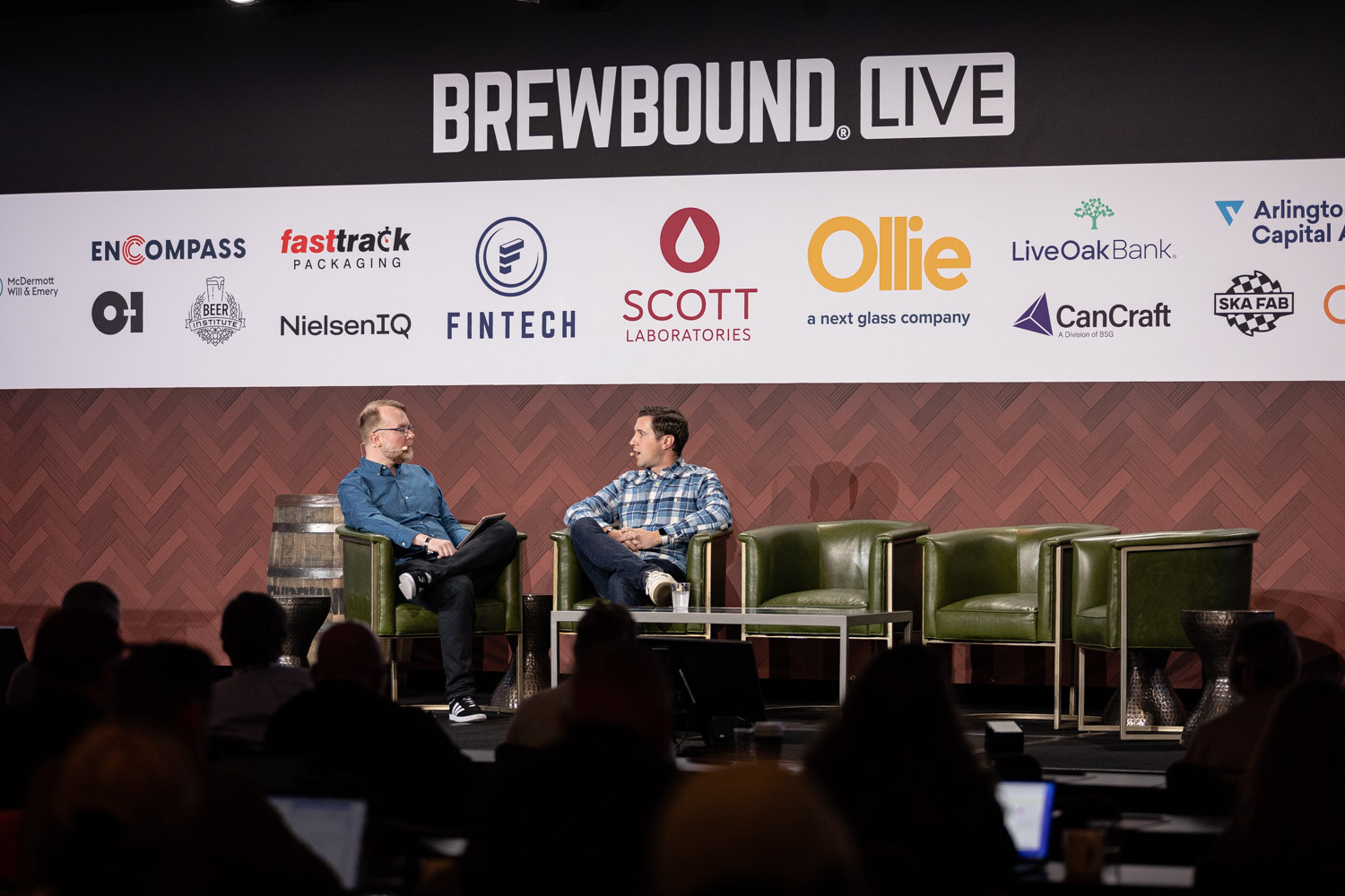Wide view of two men seated in green armchairs on a stage under a Brewbound Live banner with sponsor logos, with the dark silhouettes of the audience in front.