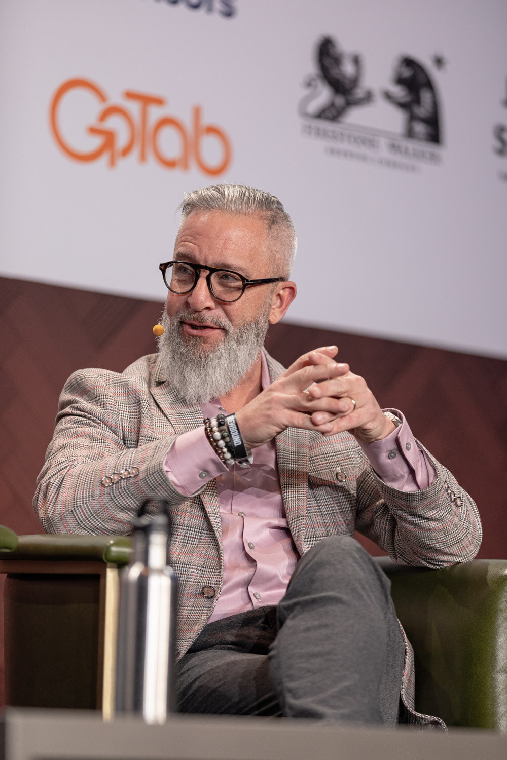 A man with a gray beard, round glasses and a plaid blazer sits in a green armchair, hands clasped, mid-sentence.