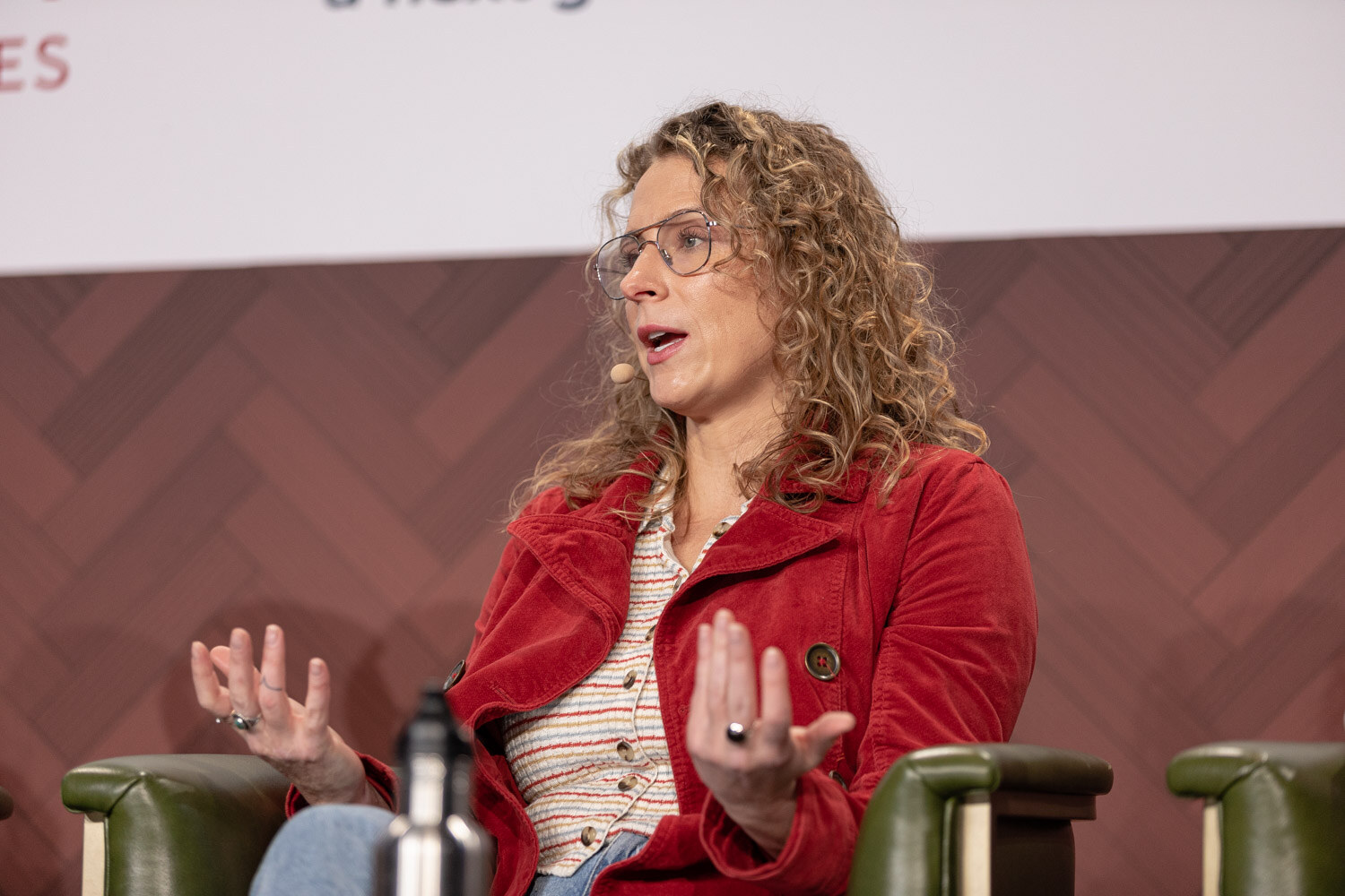 A woman with curly hair and glasses in a red jacket speaks with an open hand while seated in a green armchair.