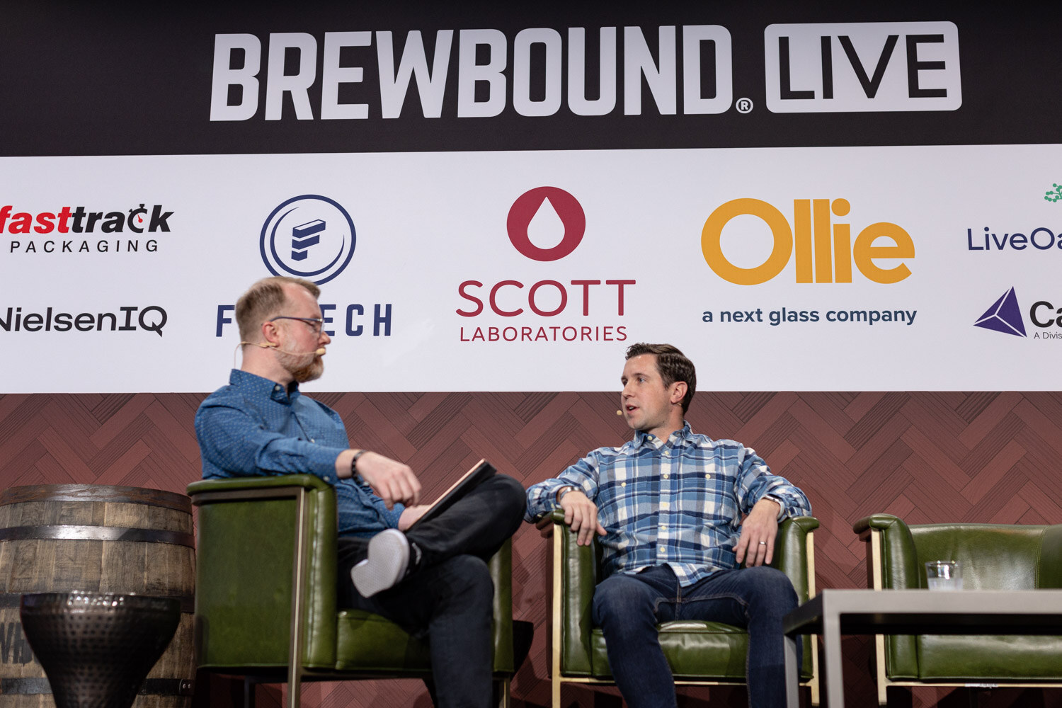 Two men in armchairs on a stage in front of a banner reading Brewbound Live and logos for Fasttrack Packaging, Scott Laboratories and Ollie.
