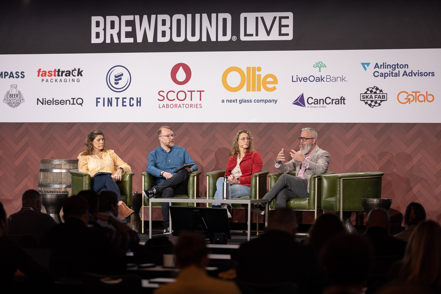 Wide view of a moderator and three panelists seated on stage under a Brewbound Live banner, with dark silhouettes of the audience in front.