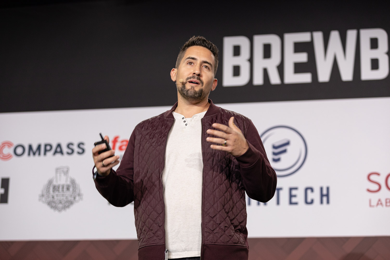 A man with short dark hair and a beard in a maroon quilted jacket and white shirt gestures in front of the Brewbound Live sponsor wall.