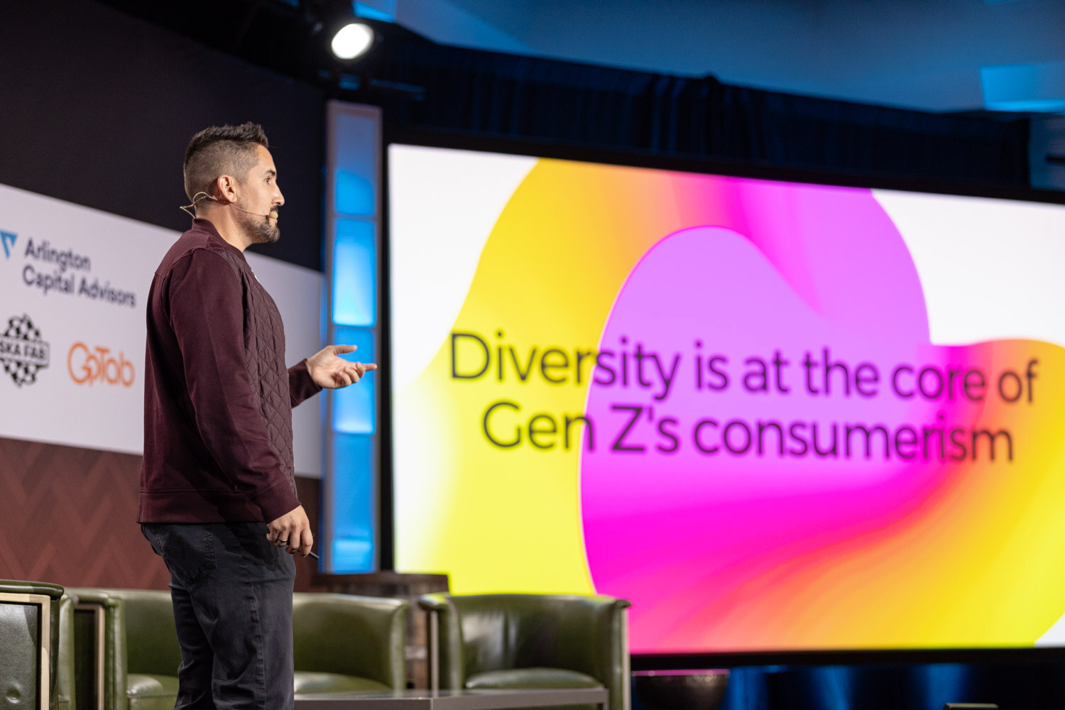 A man in a maroon jacket stands at the edge of the stage next to a bright pink and yellow slide with the words Diversity is at the core of Gen Z's consumerism.