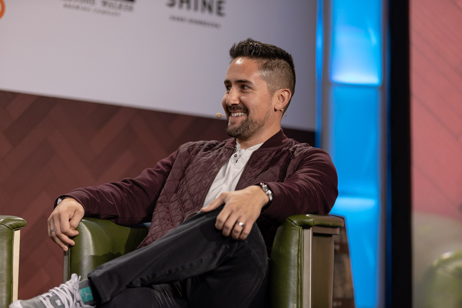 A man in a maroon quilted jacket sits in a green armchair, legs crossed, smiling off to the side.