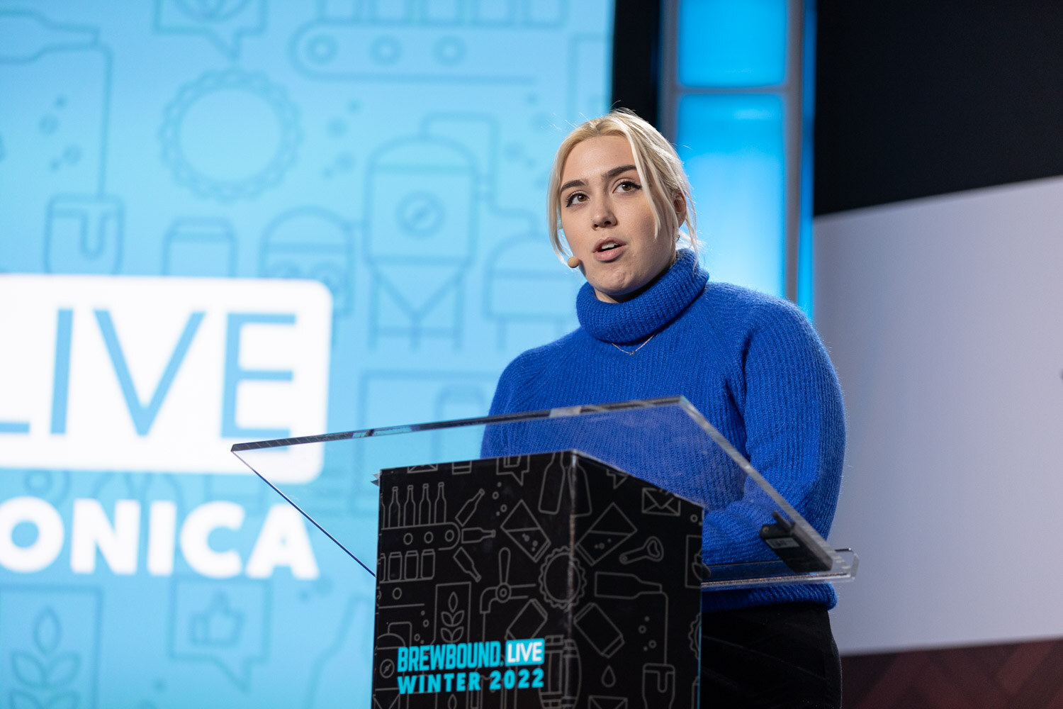 A woman with blonde hair in a blue turtleneck speaks at a clear podium in front of a blue Brewbound Live Santa Monica backdrop.