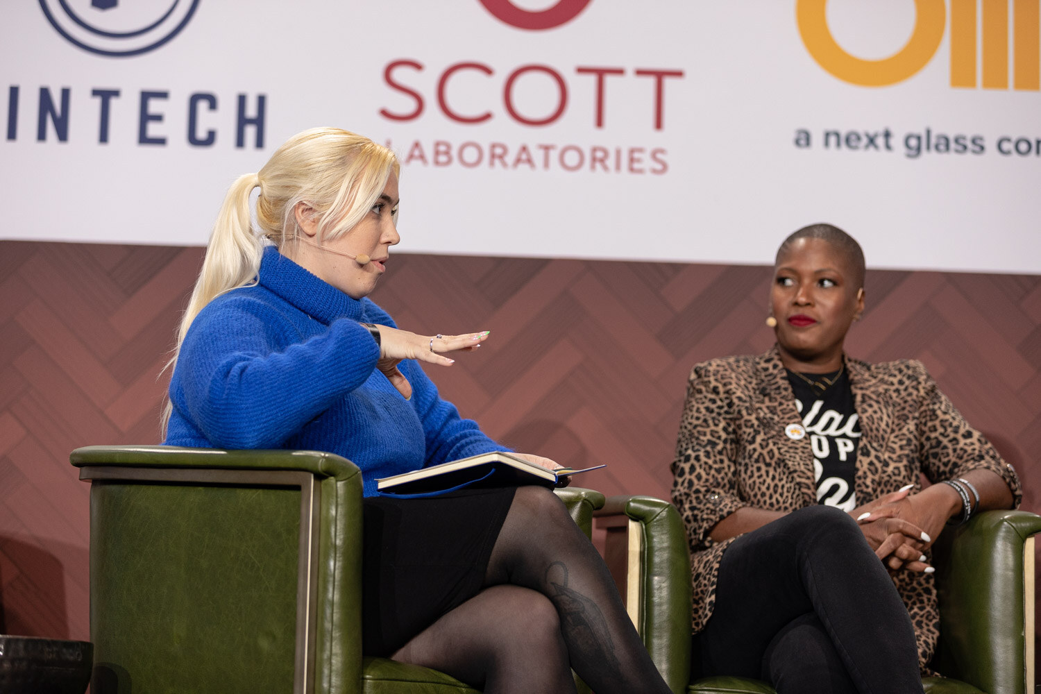 A woman with a blonde ponytail in a blue turtleneck holds a notebook and gestures toward a woman in a leopard-print blazer and black graphic T-shirt, seated in green armchairs.