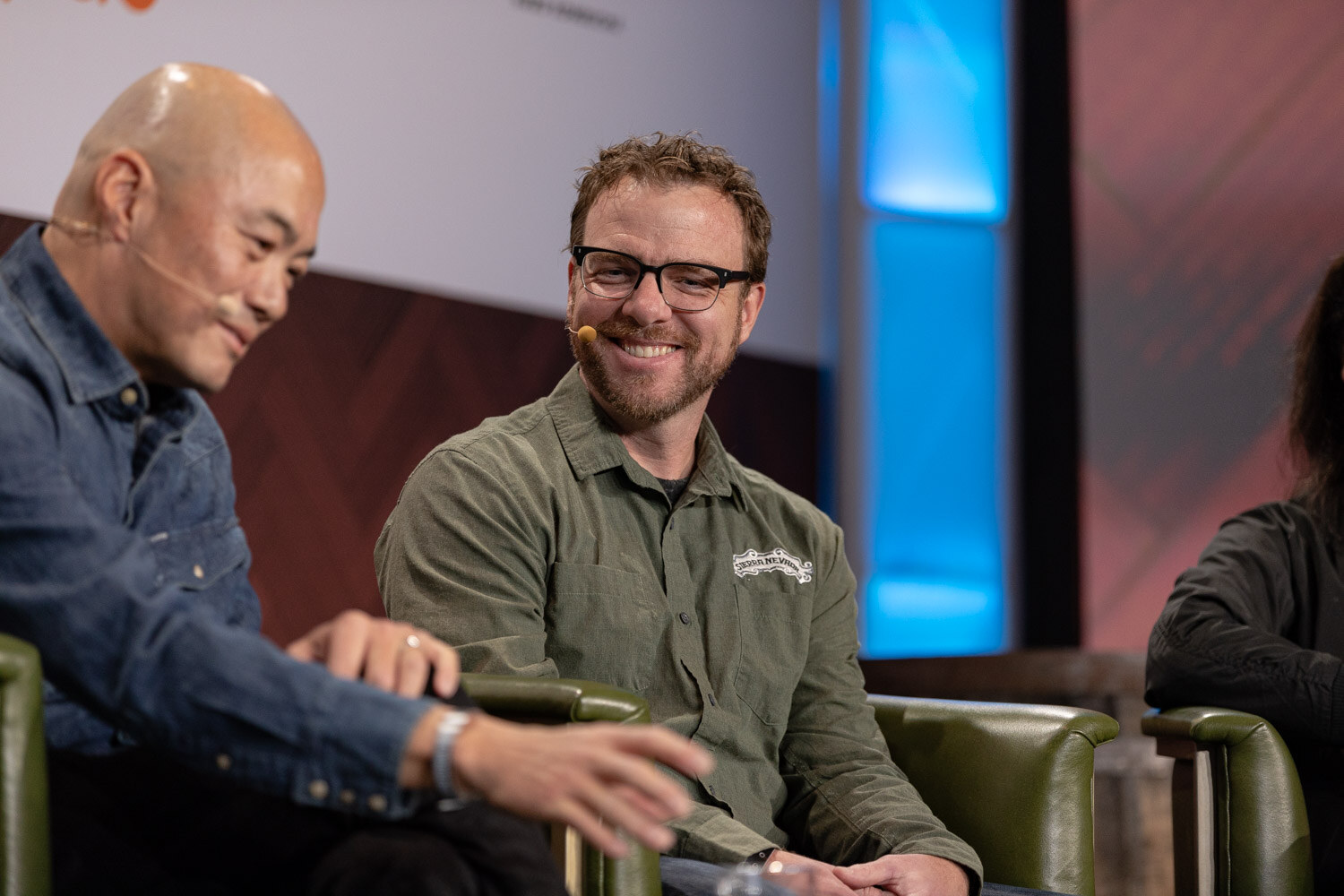A man in a denim shirt and a bearded man in a green work shirt and glasses smile at each other in green armchairs.