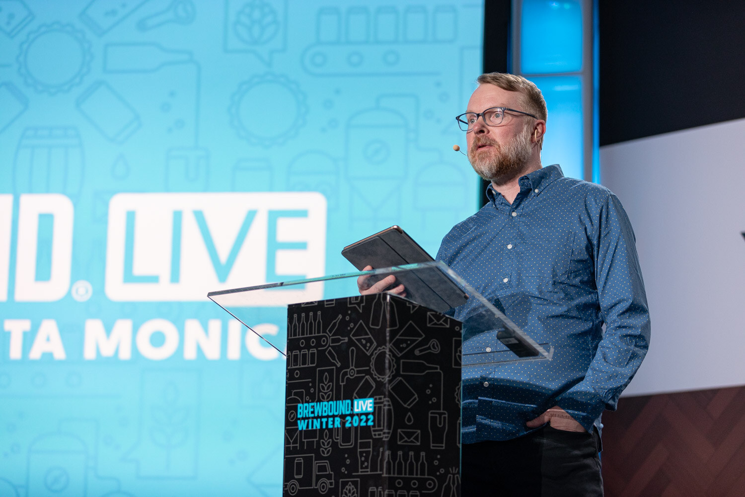 A bearded man with glasses and a headset mic in a blue button-up reads from a tablet at a clear podium in front of a Santa Monica Brewbound Live backdrop.