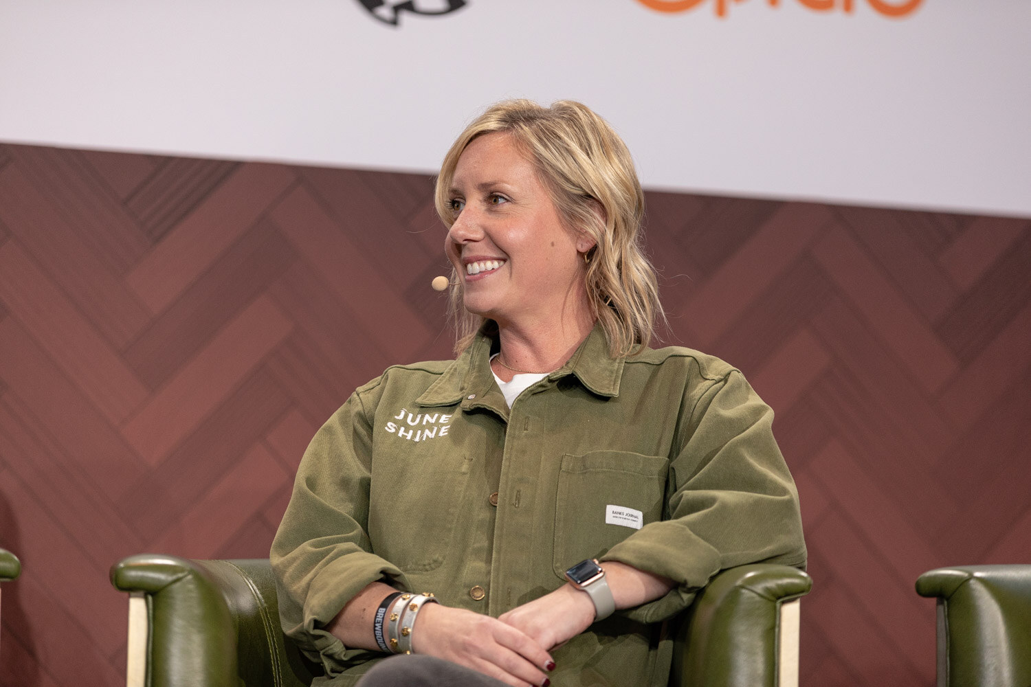 A woman with wavy blonde hair in an olive jacket printed with June Shine smiles while seated in a green armchair.