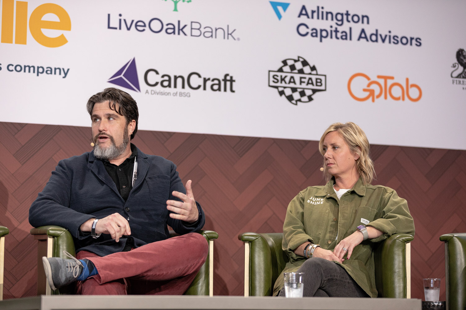 A man with a gray beard in a navy jacket and red pants gestures while a blonde woman in an olive jacket sits beside him in a green armchair.