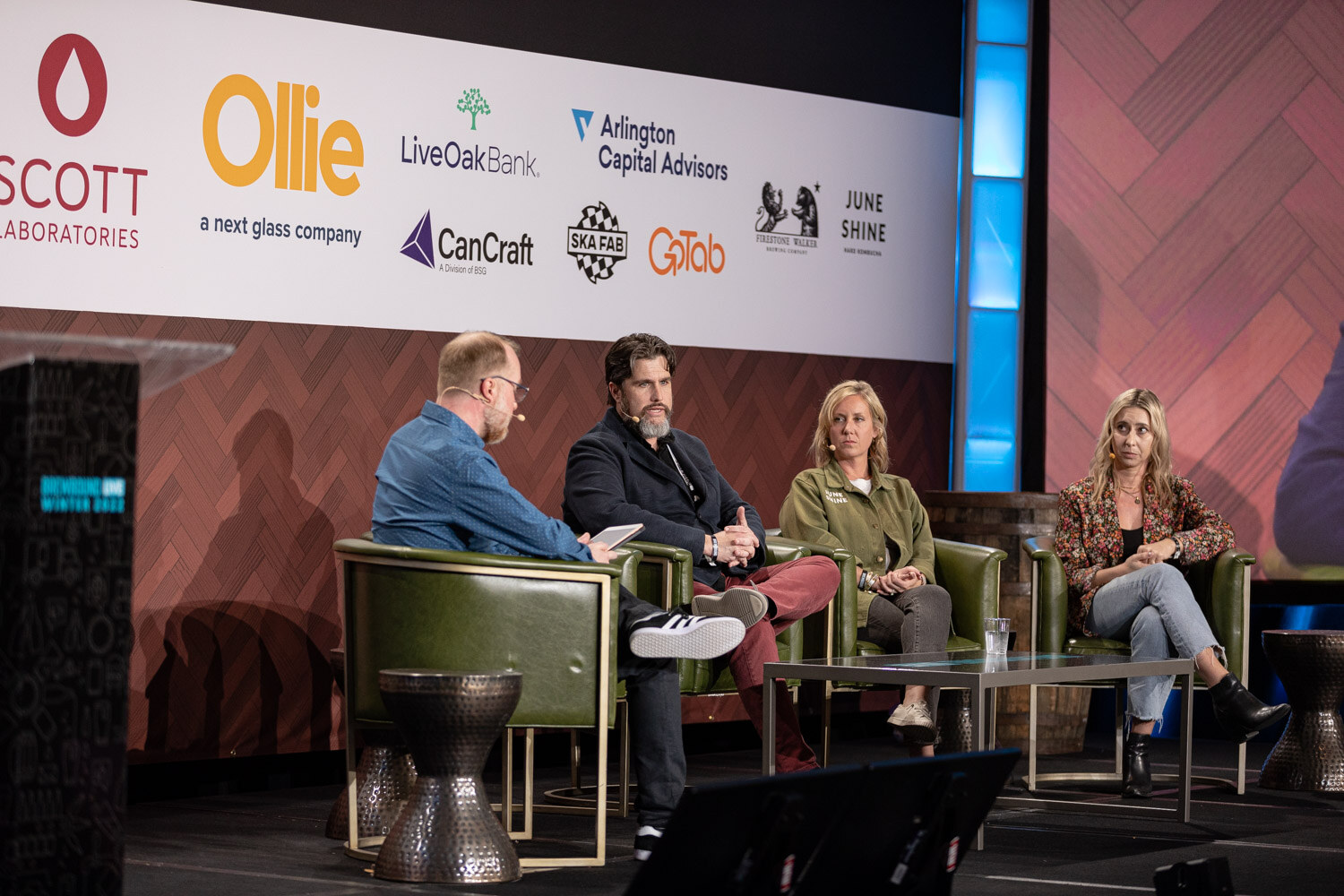 Wide view of four people seated in green armchairs on a stage under the Ollie, Scott Laboratories and CanCraft sponsor backdrop.