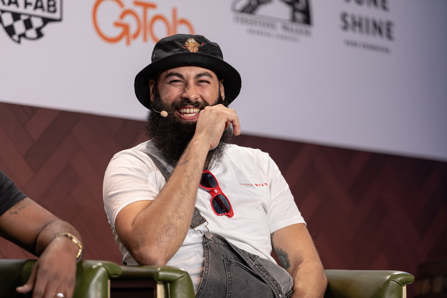 A smiling bearded man in a black bucket hat and white shirt with sunglasses hooked on his collar laughs on stage.