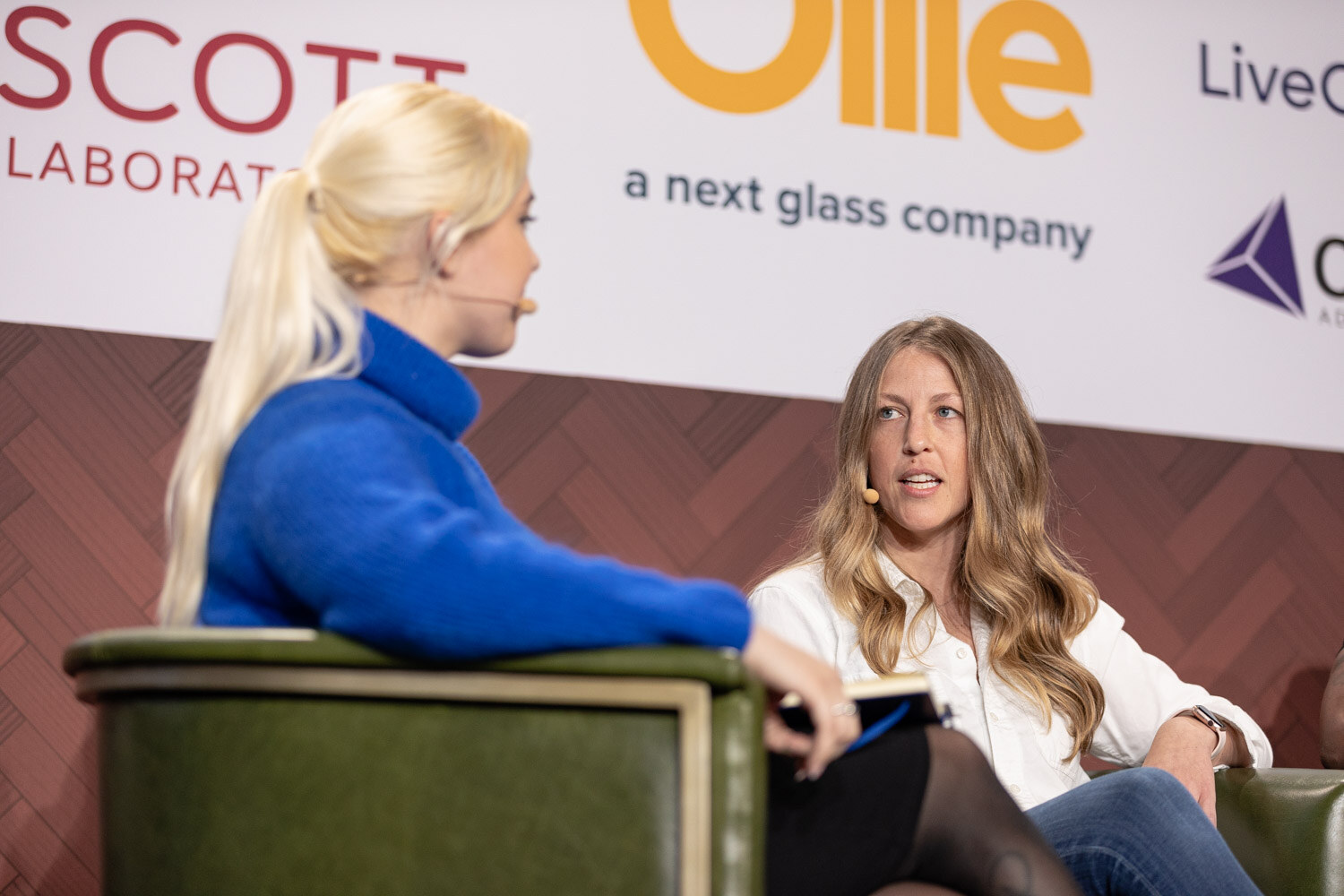 A woman with a blonde ponytail in a blue turtleneck faces a woman with long wavy hair in a white blouse, both seated on stage.