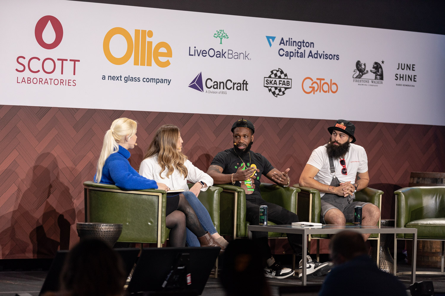 Wide view of four people in green armchairs on a stage in front of a sponsor wall with Scott Laboratories, Ollie and CanCraft logos.