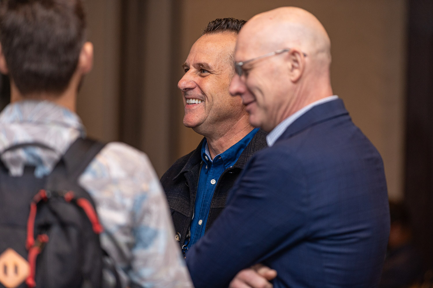 A smiling man in a blue shirt and a bald man in a navy blazer talk, with a person in a backpack in the foreground.