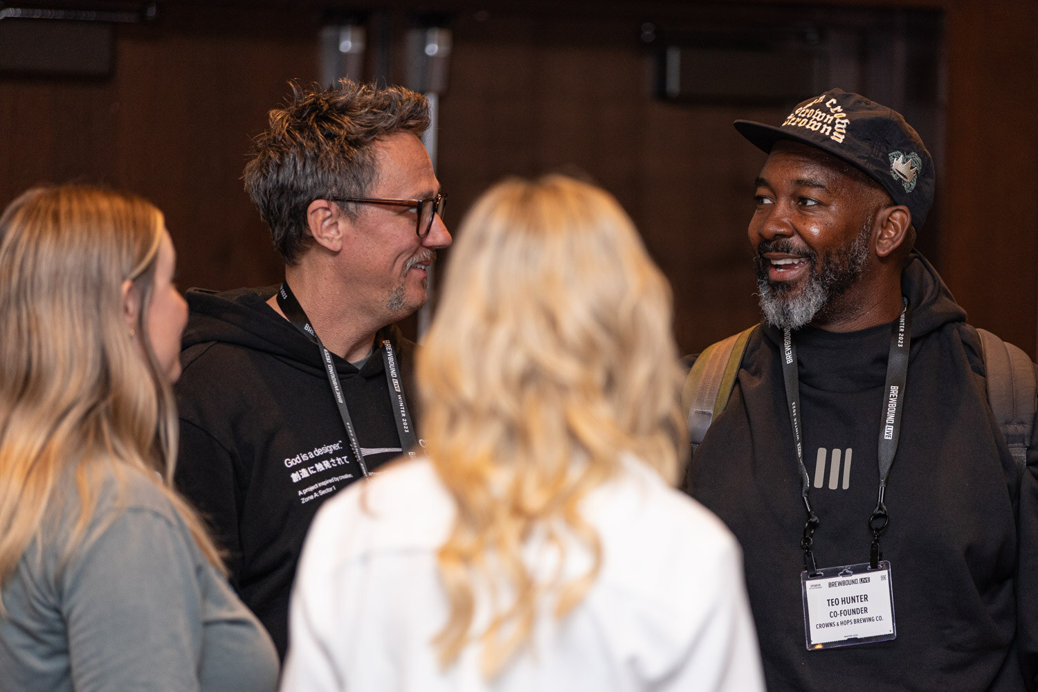 A bearded man in a black cap with a lanyard smiles at a man in a black hoodie, with two women with blonde hair in the foreground.