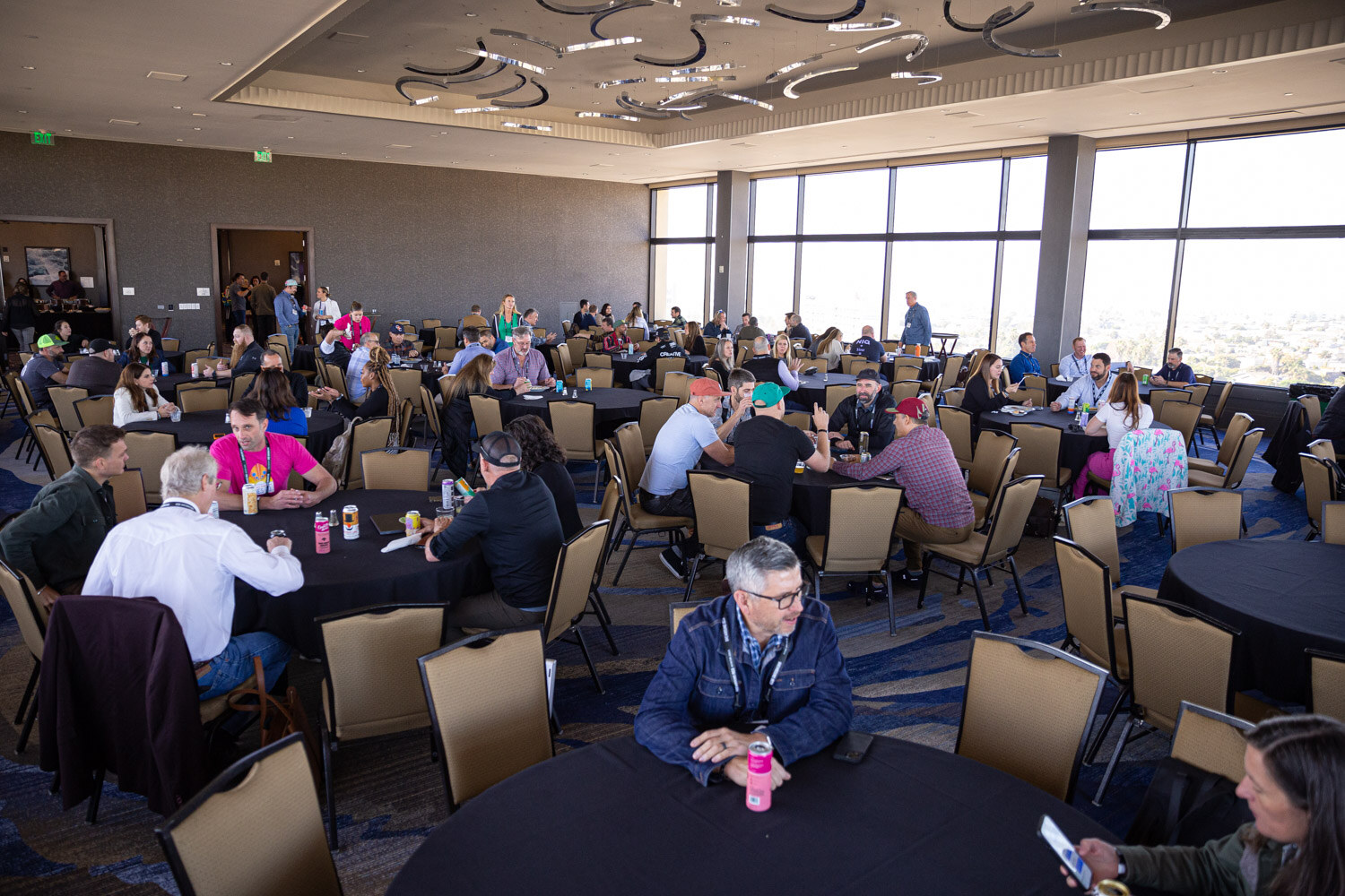 A wide view of a ballroom with round black tables and attendees seated at lunch, with large windows in the background.
