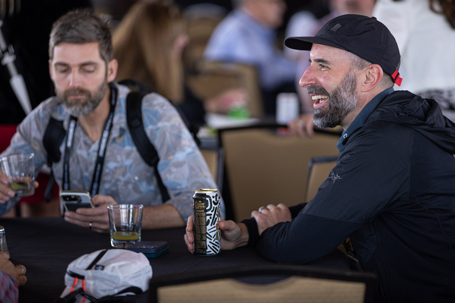 A man in a patterned shirt looks at his phone while a smiling bearded man in a black cap sits beside him at a table with a can.