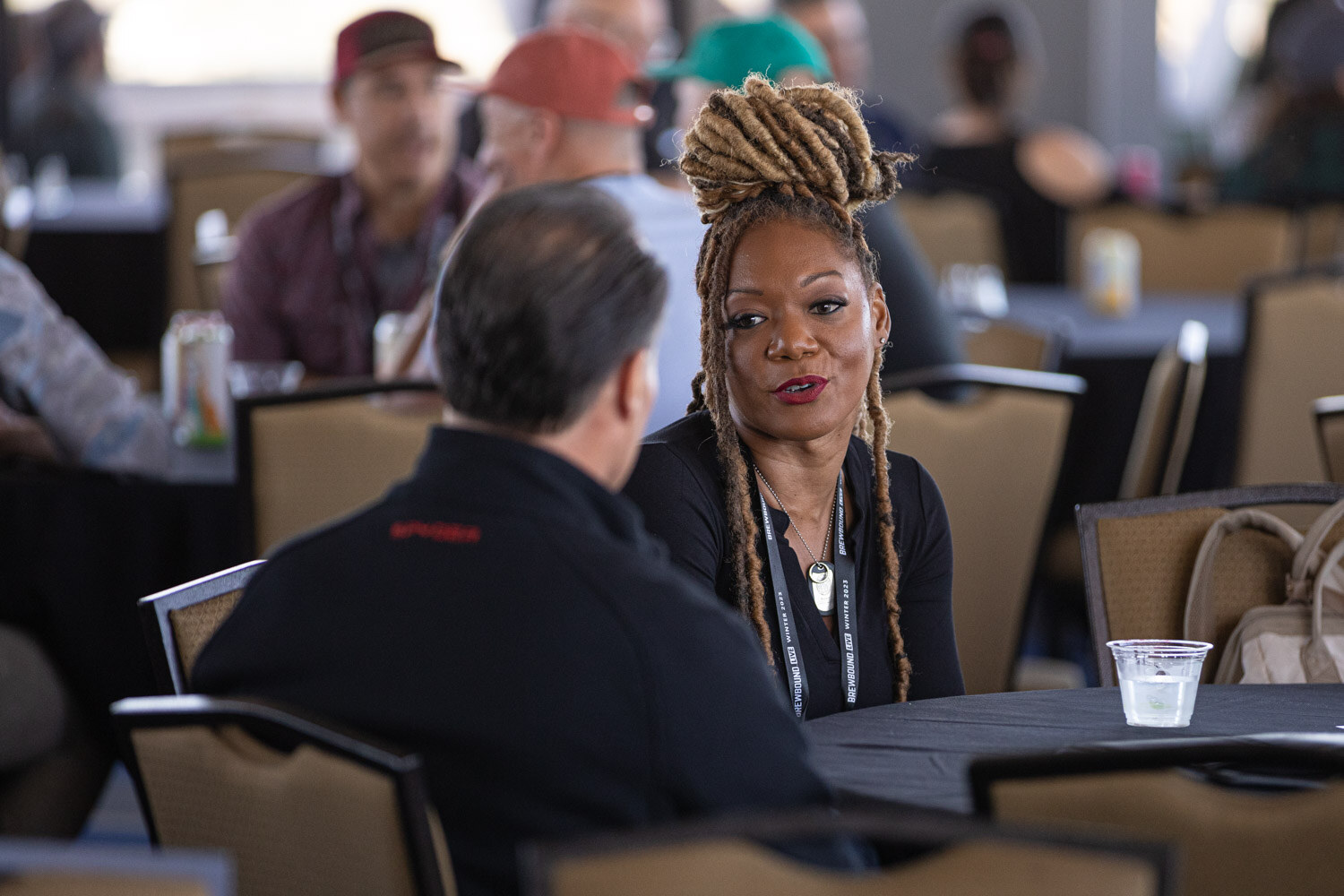 A woman with braided hair in a black top speaks with a man seen from behind at a round table.