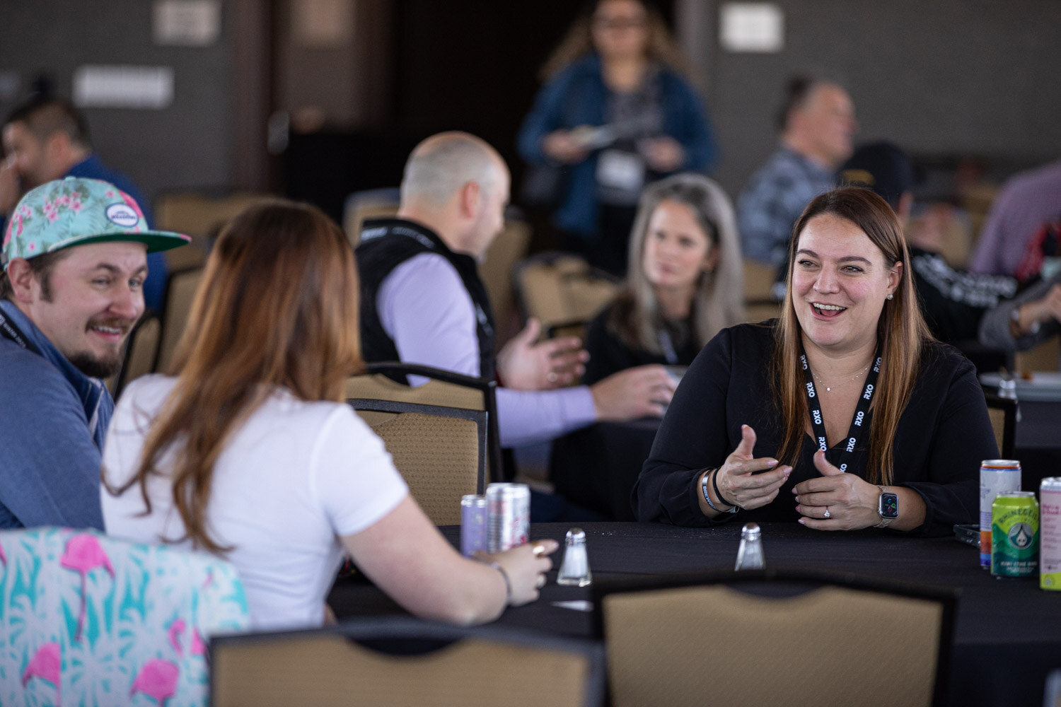A woman in black laughs and gestures while attendees sit around a table with cans.