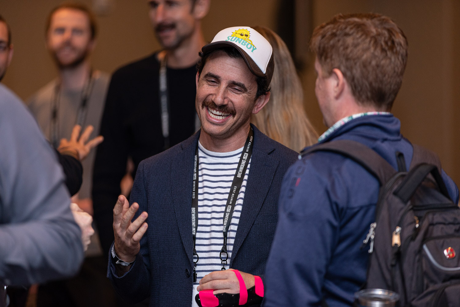 A man in a trucker cap, striped shirt and navy blazer smiles and gestures while talking with others.