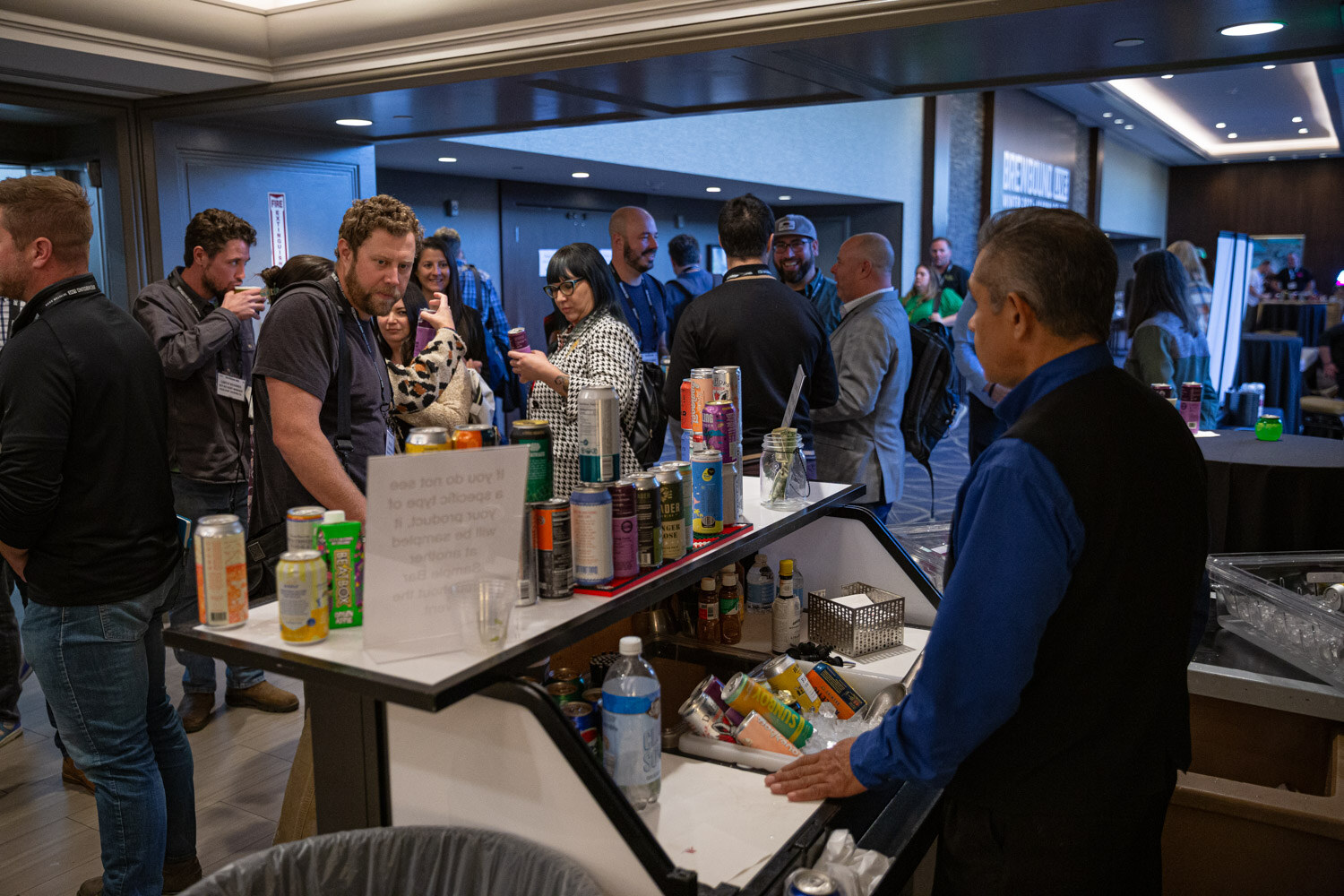 A crowd of attendees stands around a cart holding many cans while a man in a vest tends it.