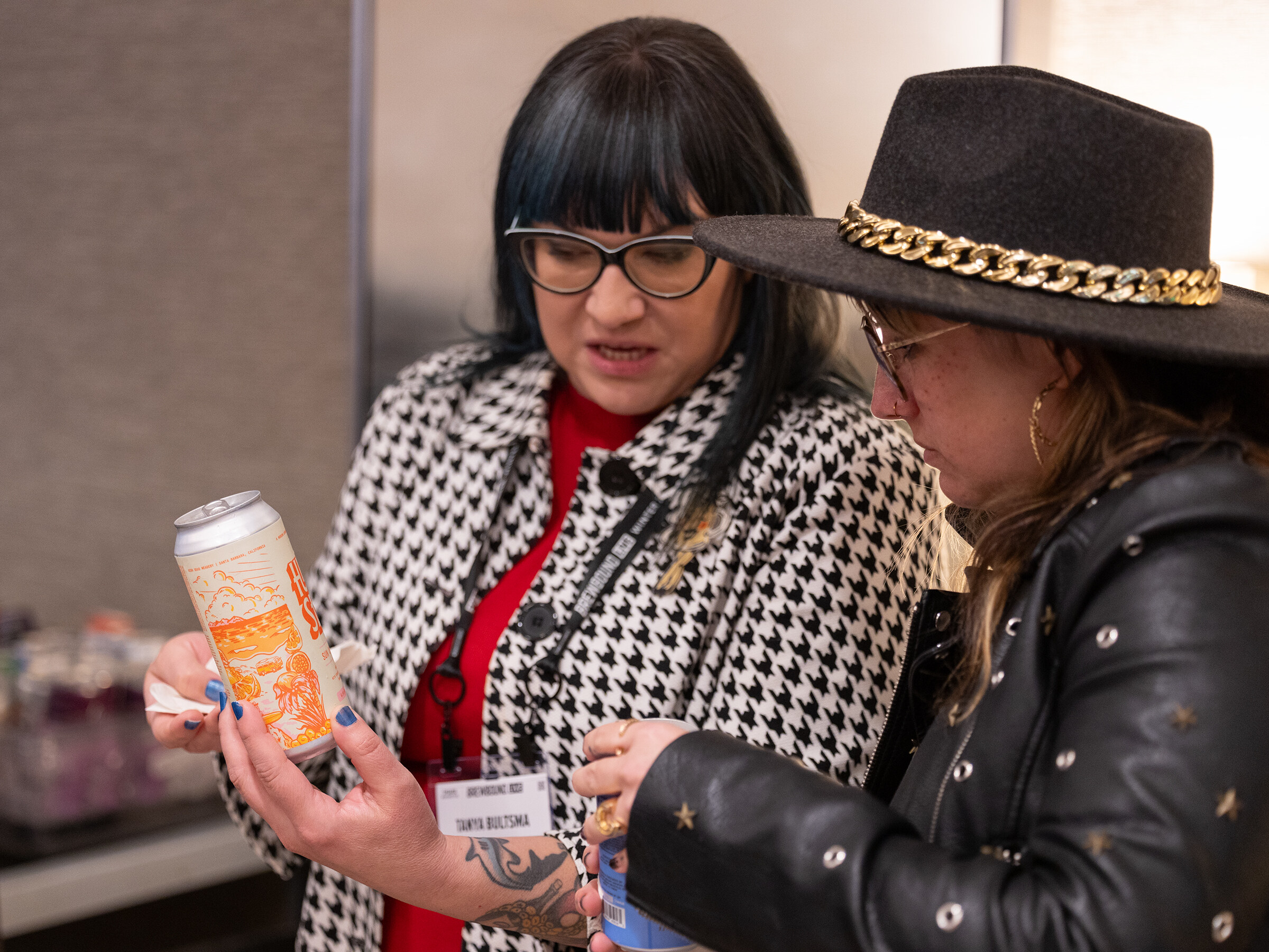 A woman in a houndstooth jacket holds up an orange can while a woman in a black hat with a chain band looks on.
