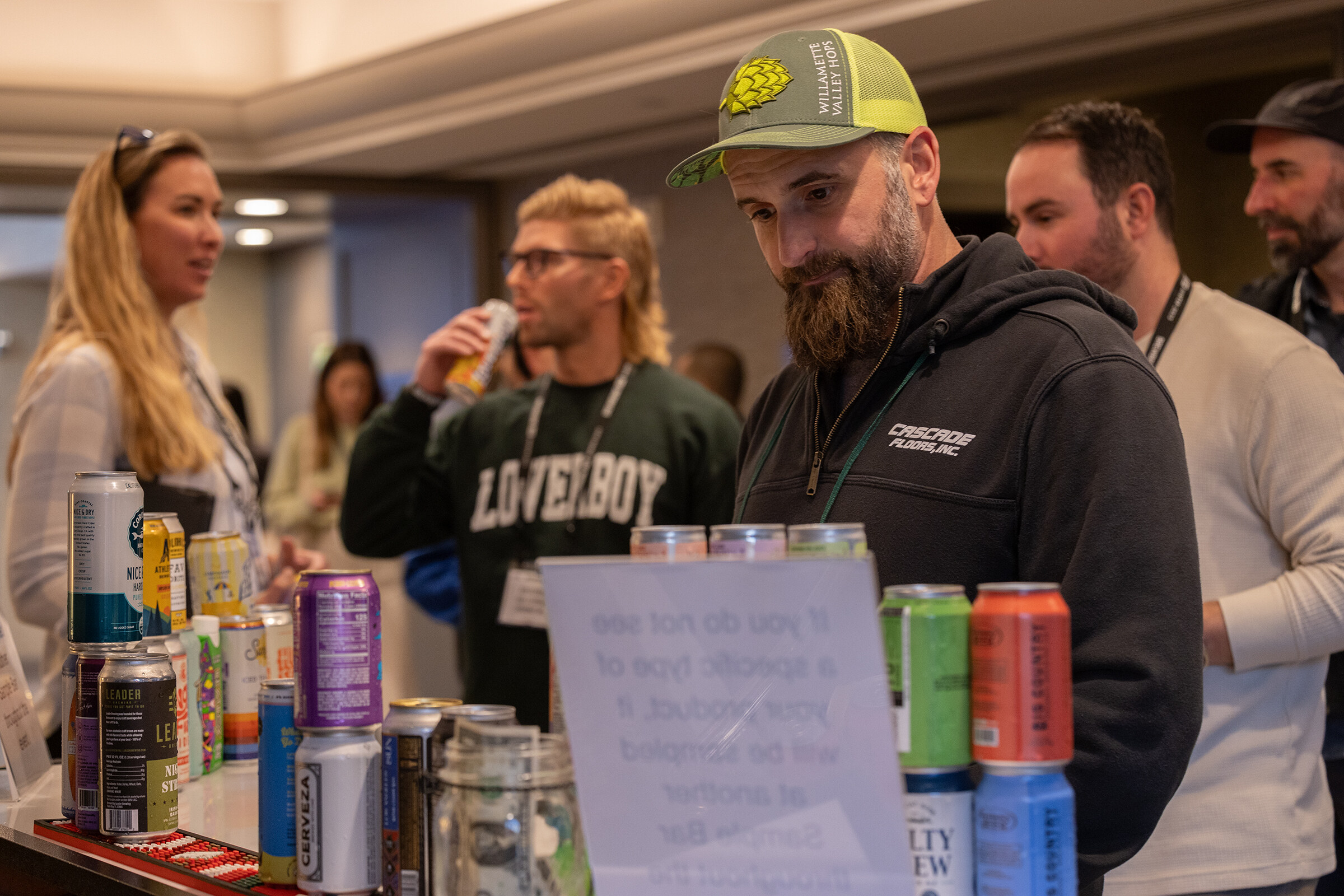 A bearded man in a hop-logo cap looks over a table of assorted cans while other attendees stand nearby.