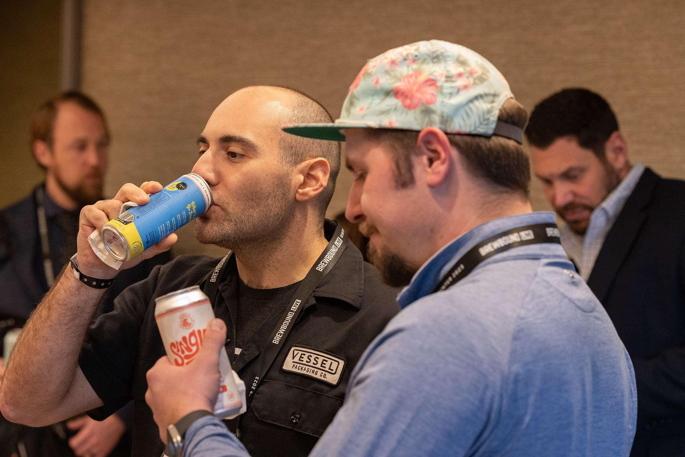 A man in a black shirt drinks from a blue and yellow can while a man in a floral cap holds another can beside him.
