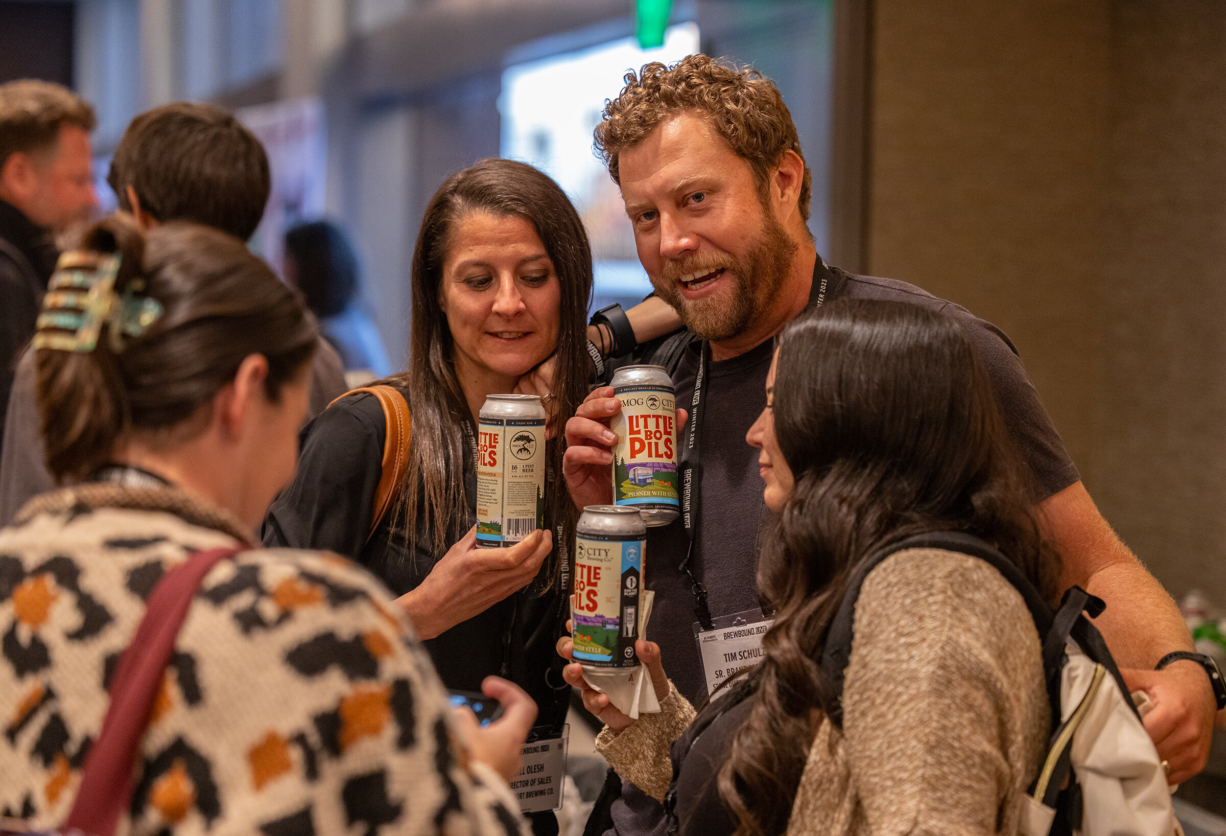 A bearded man holds up a can of Little Pils while several women and men talk around him.