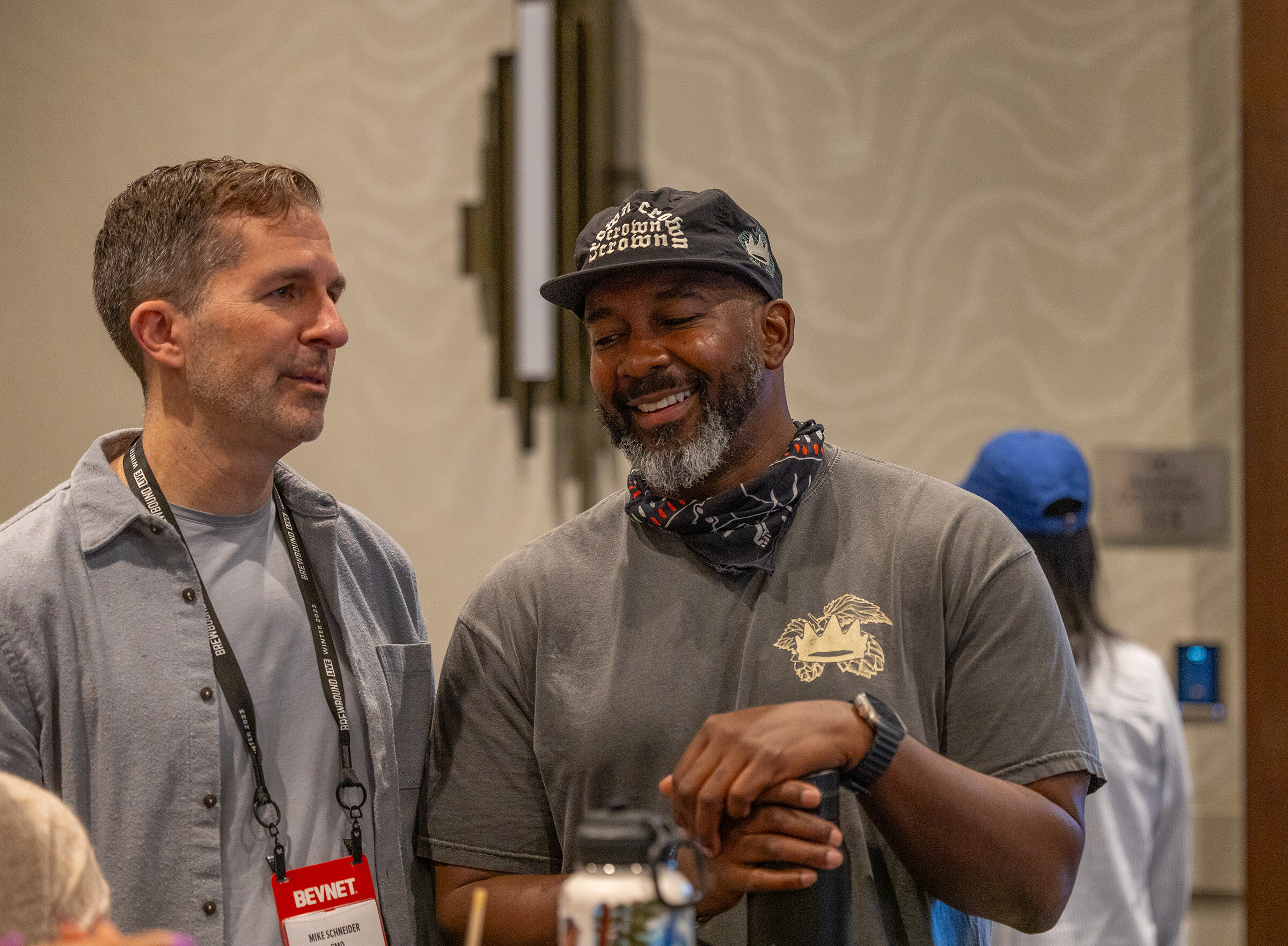 A man in a gray shirt and a bearded man in a gray t-shirt and bandana laugh together, both wearing lanyards.