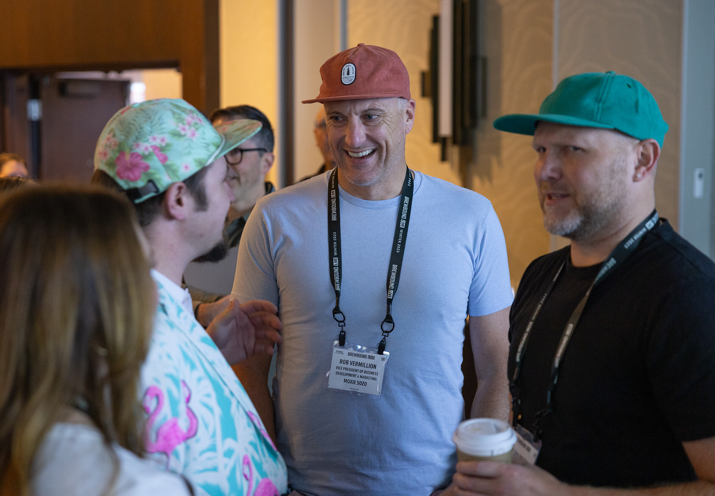 Three men in caps and lanyards, one in a bright flamingo-print jacket, laugh together.