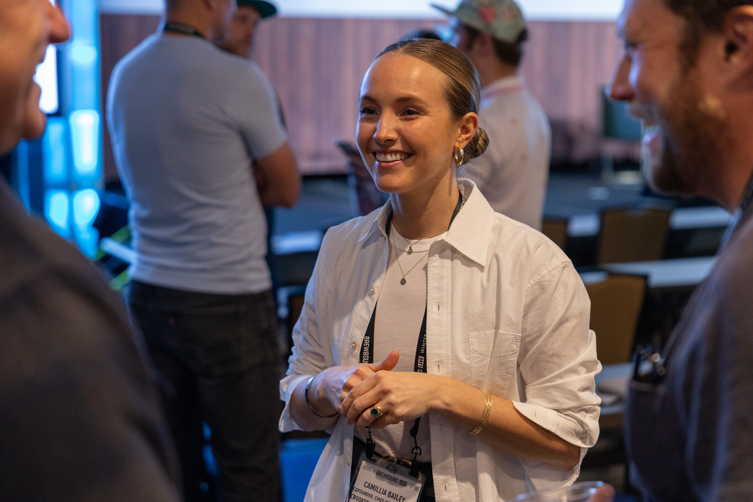 A smiling woman in a white button-up shirt with a lanyard stands among other attendees.