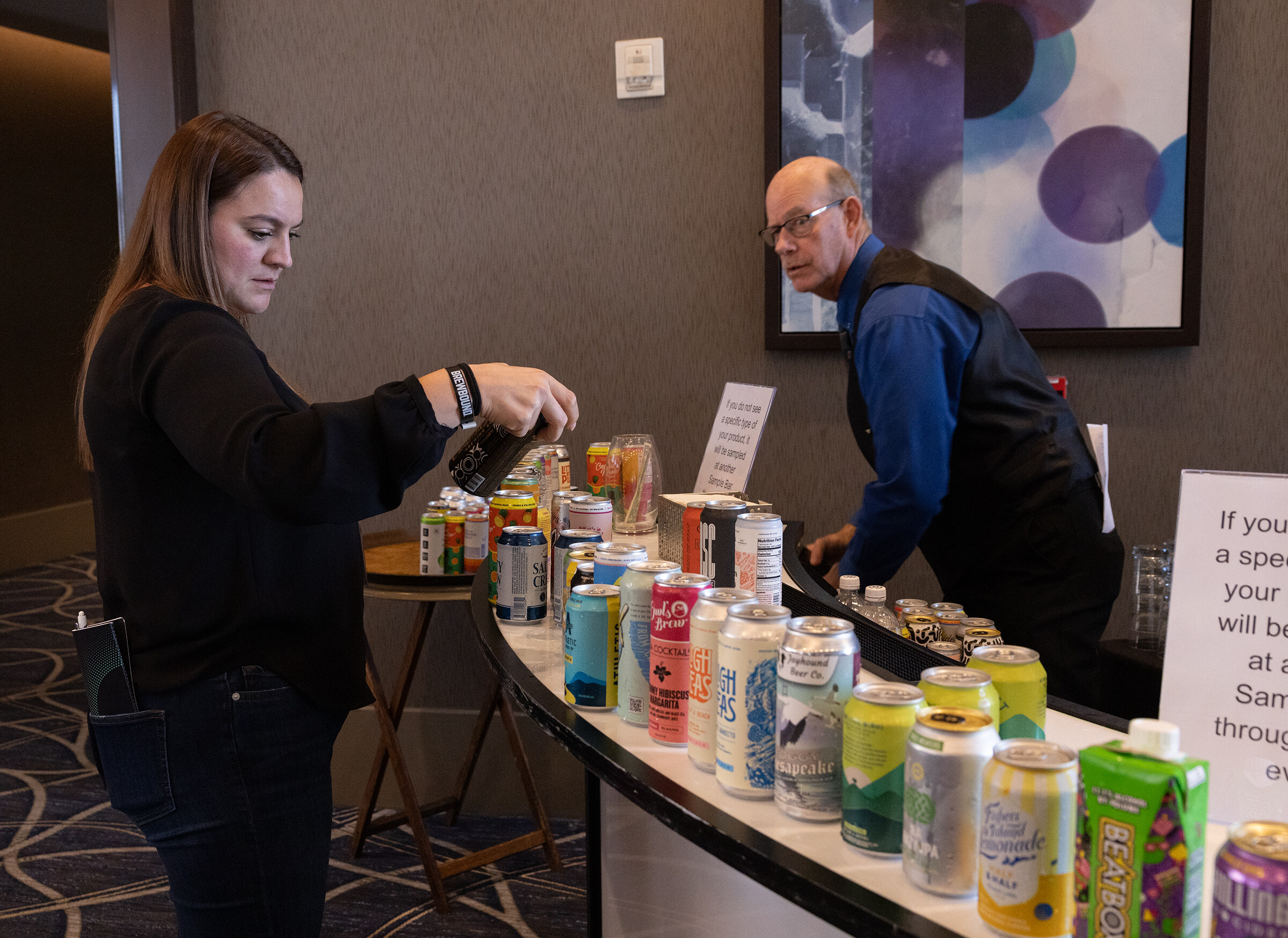 A woman in black picks up a can from a curved bar covered in beverage cans while a man in a vest stands behind it.