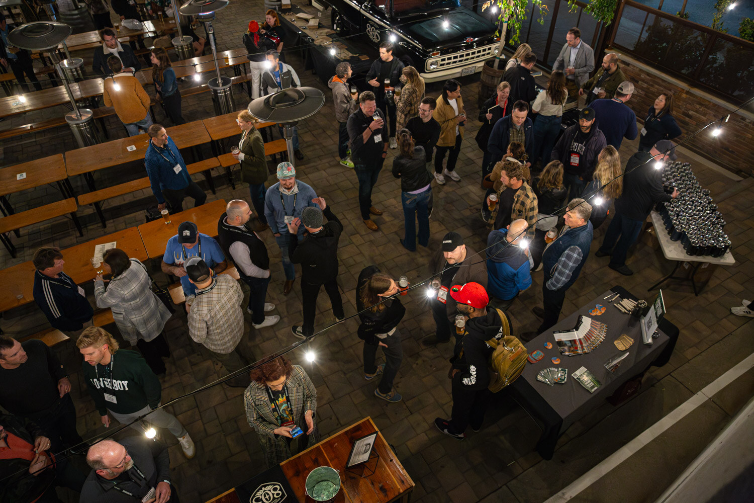 An overhead view of guests standing and sitting on a lit brewery patio with long wooden tables and a vintage truck.